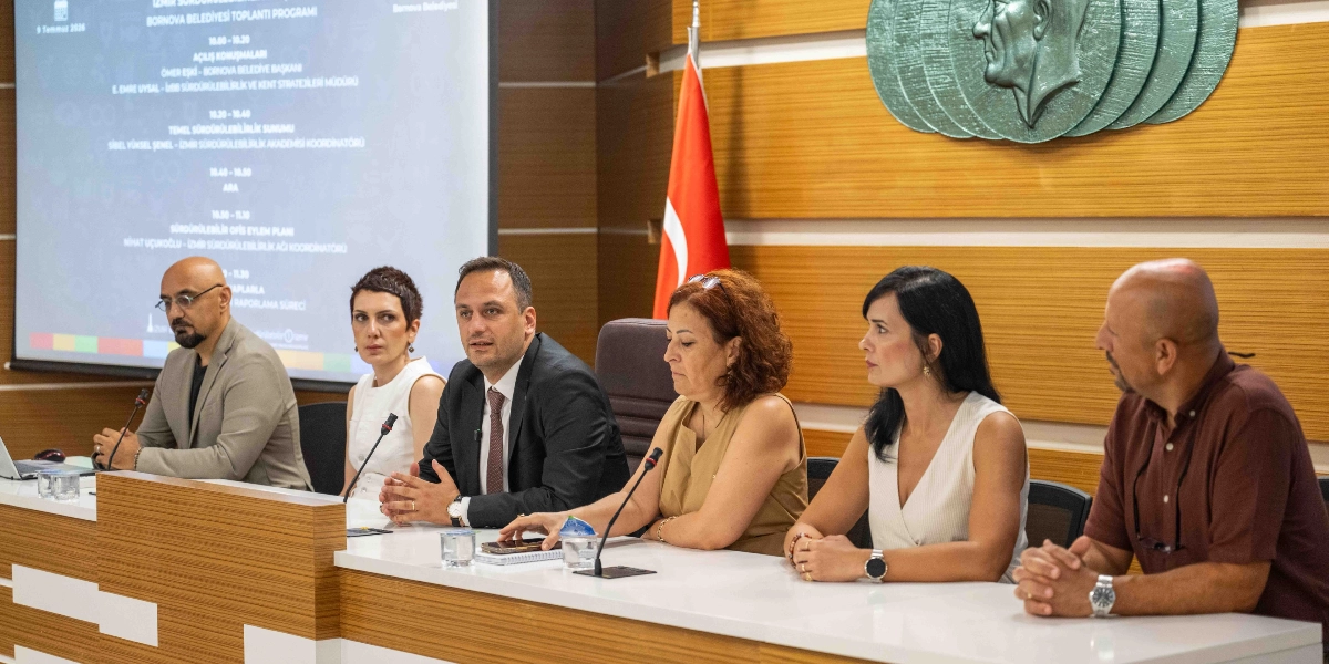 Bornova Belediyesi'nde düzenlenen bir toplantıda, uzun bir masa etrafında yan yana oturan altı kişilik bir panel grubu yer alıyor. Masanın ortasında konuşma yapan Bornova Belediye Başkanı Ömer Eşi ve yanındaki uzmanlar, sürdürülebilirlik konulu bir sunum gerçekleştiriyor. Arka planda, üzerinde toplantı programının detaylarının yer aldığı büyük bir projeksiyon ekranı ve duvarda Atatürk silüetinin bulunduğu kabartmalı bir pano ile Türk bayrağı görülüyor.