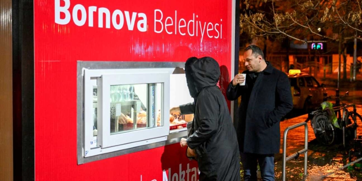 Üzerinde Bornova Belediyesi yazan kırmızı renkli bir yardım standının önünde, gece vakti bir vatandaş pencereden yiyecek alırken, yanında duran bir erkek elinde sıcak içecek bardağıyla bekliyor.