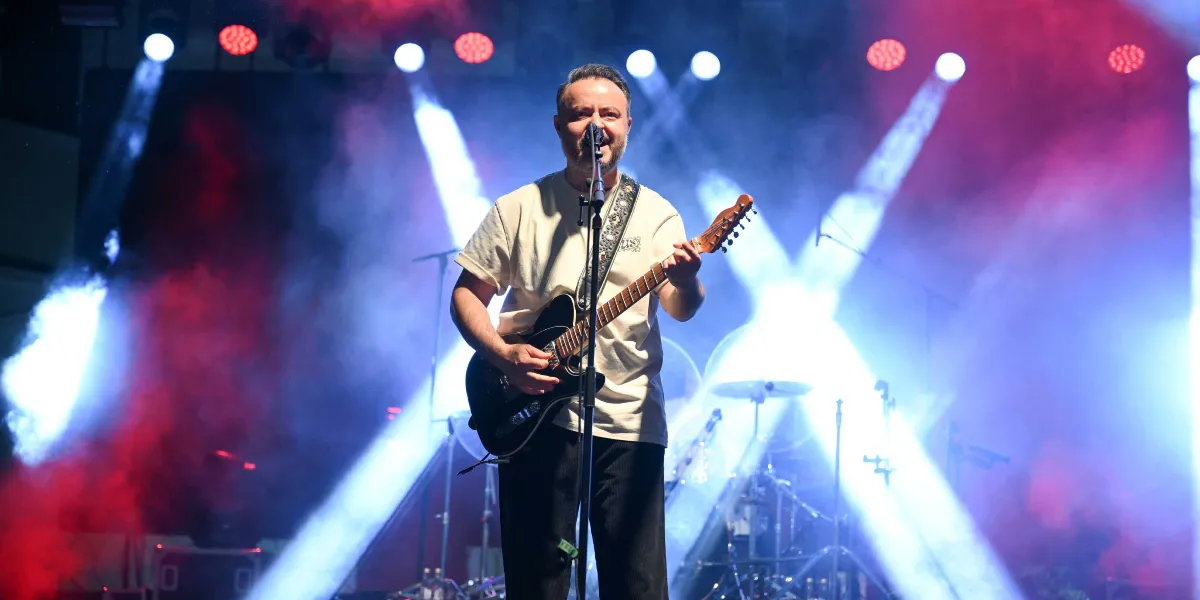 Sahne ışıkları ve yoğun sisin arasında, beyaz tişörtlü ve siyah pantolonlu bir erkek şarkıcı, elinde elektro gitarıyla mikrofon başında şarkı söylüyor. Arka planda davul seti ve sahne ekipmanları seçilirken, ortamı kırmızı ve mavi renkli güçlü spot ışıkları aydınlatıyor.