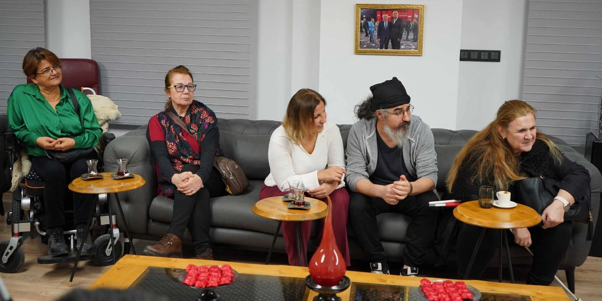 Modern bir ofis ortamında, gri bir koltukta yan yana oturan beş kişilik bir grup. En solda tekerlekli sandalyede oturan yeşil gömlekli bir kadın, yanında bordo üstlü ve desenli şallı bir kadın, beyaz bluzlu bir kadın, başında siyah bir bere olan sakallı bir erkek ve en sağda siyah giyimli uzun saçlı bir kadın yer alıyor. Önlerinde ahşap sehpalar üzerinde çay bardakları ve kırmızı tatlılar bulunuyor. Arka planda duvarda çerçeveli bir fotoğraf asılı.