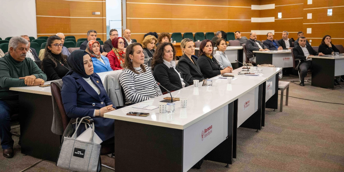 Bornova Belediyesi'nin düzenlediği bir toplantıda, uzun beyaz masaların etrafında yan yana oturan, aralarında kadın ve erkek muhtarların bulunduğu bir grup katılımcı. Katılımcılar dikkatle karşıya bakarken, masaların üzerinde Bornova Belediyesi logosu yer alıyor. Arka planda ahşap panellerle kaplı duvarlar ve düzenli bir oturma düzeni görülüyor.