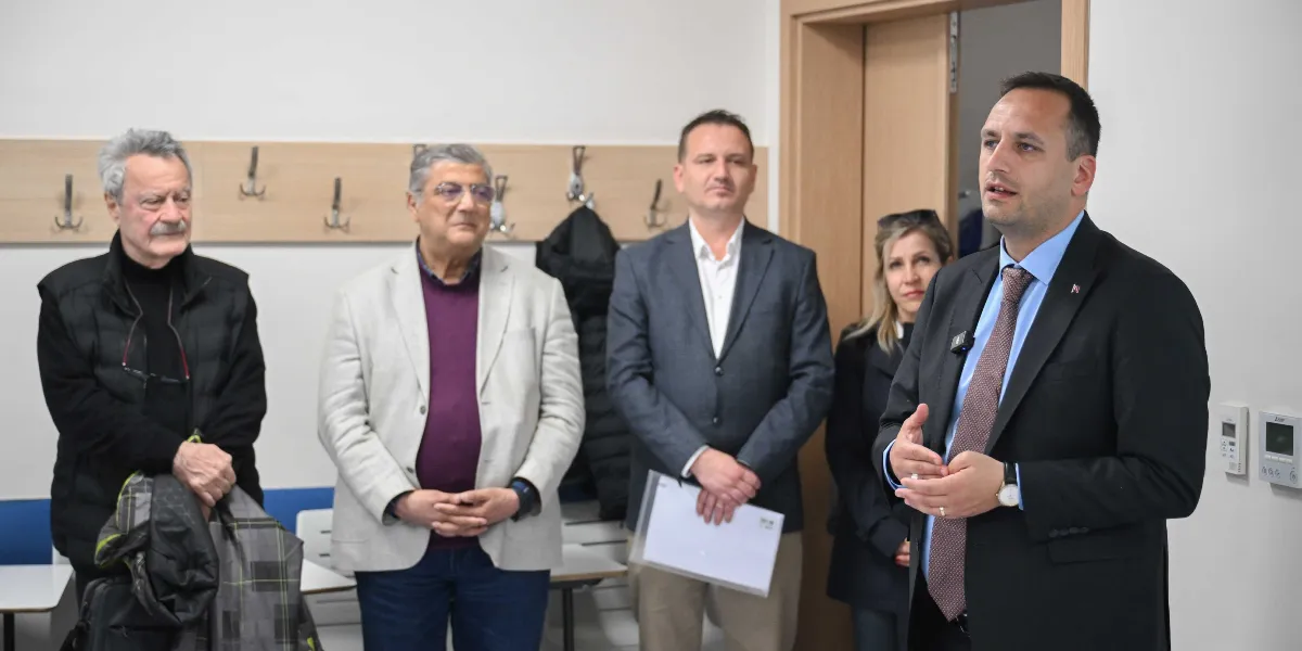 Bornova Belediye Başkanı Ömer Eşki, bir toplantı odasında ayakta durarak konuşma yapıyor. Eşki'nin yanında ve arkasında, onu dikkatle dinleyen üç erkek ve bir kadın bulunuyor. Katılımcılar resmi kıyafetler giymiş durumda ve ortamda duvarda askılıklar ile beyaz bir yazı tahtası görülüyor.