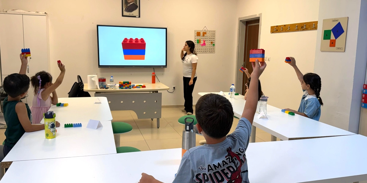 Sınıfta öğretmen, akıllı tahtada gösterilen kırmızı, mavi ve turuncu renkli üç bloktan oluşan yapıyı öğrencilere anlatıyor. Öğrenciler ellerinde tuttukları renkli plastik bloklarla öğretmeni takip ediyor ve bazıları ellerini havaya kaldırarak derse katılıyor. Masaların üzerinde su mataraları ve farklı renklerde bloklar bulunuyor.