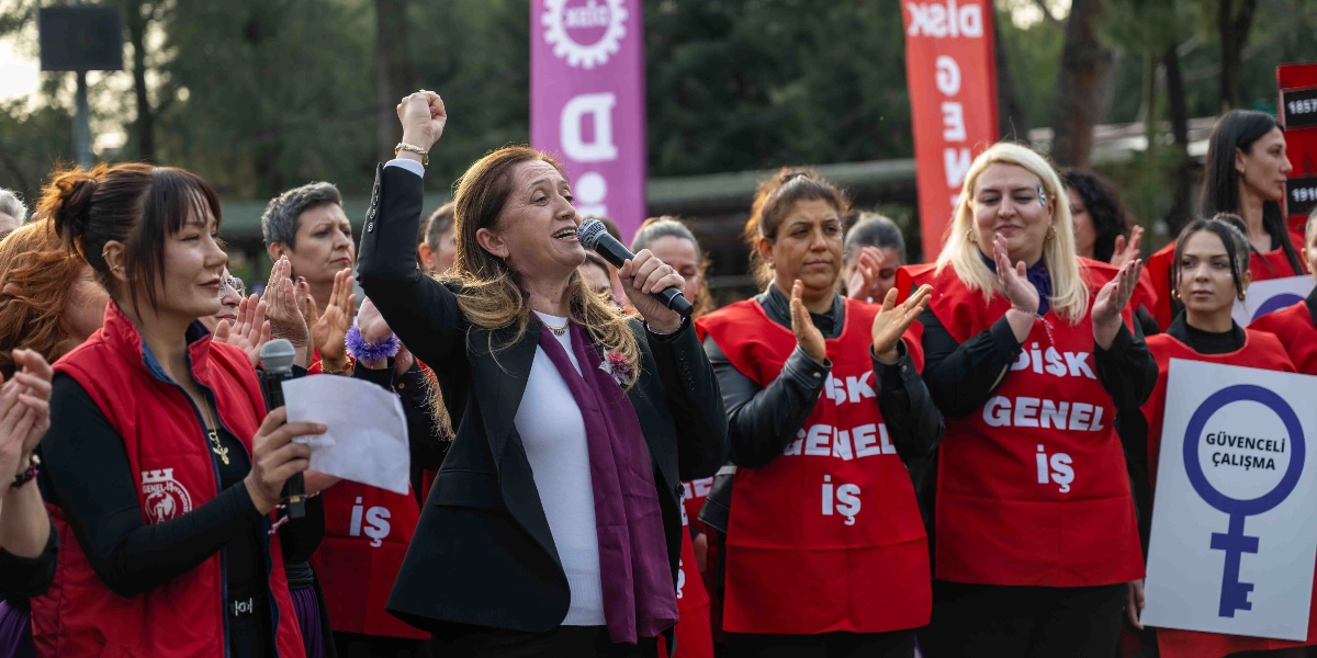 Açık havada düzenlenen bir etkinlikte, siyah ceketli bir kadın elinde mikrofonla konuşma yaparken sağ elini havaya kaldırmış durumda. Etrafındaki kadınların çoğu üzerinde 
