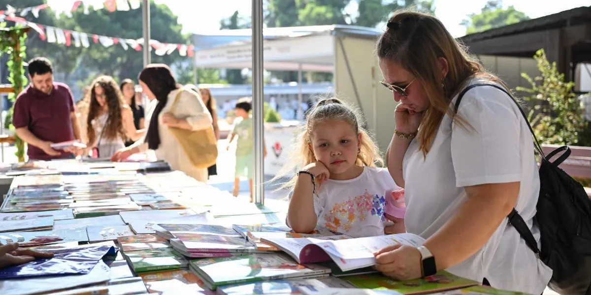 Açık havada kurulan bir kitap fuarı standında, beyaz tişörtlü bir kadın ve yanındaki küçük kız çocuğu masanın üzerindeki kitapları inceliyor. Arka planda kitaplara bakan diğer ziyaretçiler ve gölgelikli stantlar görülüyor.