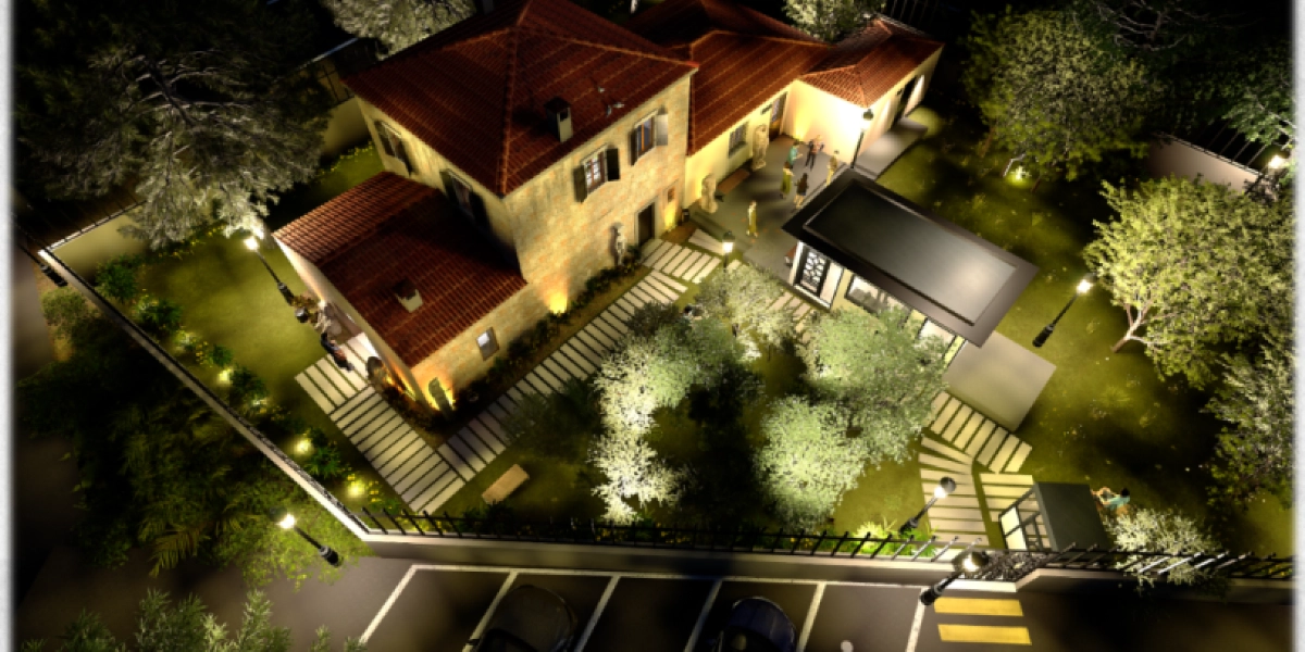 Gece aydınlatılmış, kiremit çatılı taş mimariye sahip Girit Anı Evi'nin kuş bakışı görünümü. Bahçesinde yürüyüş yolları, ağaçlar ve aydınlatma direkleri bulunan yapının önünde ziyaretçiler yer alıyor. Binanın yanında modern tasarımlı bir ek yapı ve alt kısımda park alanı bulunuyor.
