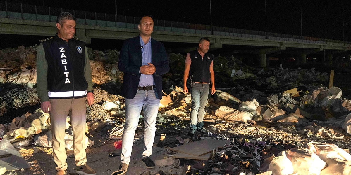 Gece vakti, arkada bir viyadüğün bulunduğu açık arazide, yığılmış moloz ve atıkların önünde duran üç erkek. Solda üzerinde 