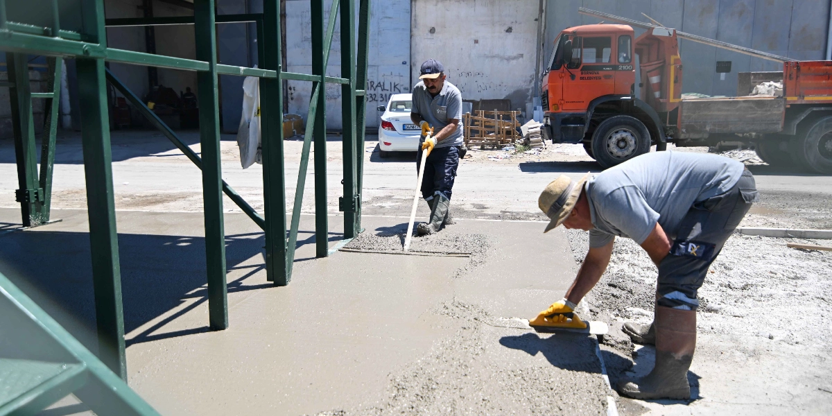 İki işçi, Bornova Belediyesi'ne ait bir kamyonun yakınında, yeni dökülen beton zemini düzeltmek için çalışıyor. İşçilerden biri elindeki uzun saplı aletle betonu yayarken, diğeri el malasıyla yüzeyi pürüzsüzleştiriyor. Arka planda beyaz bir binek otomobil ve inşaat alanı görülüyor.