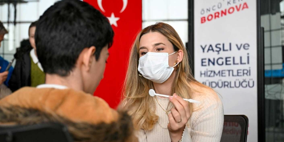 Beyaz maskeli bir diş hekimi, karşısında oturan bir öğrenciye elindeki diş aynasıyla ağız ve diş sağlığı taraması yapıyor. Arka planda üzerinde 