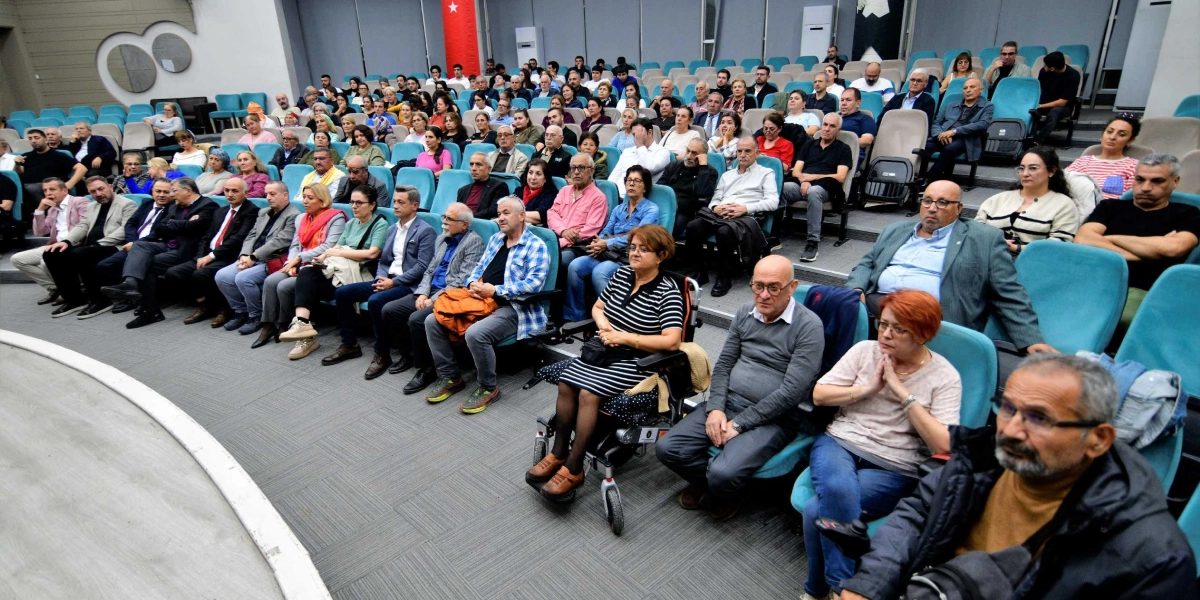Modern bir konferans salonunda, Tahtacı Türkmen kültürü üzerine düzenlenen bir etkinliği izleyen kalabalık bir dinleyici grubu. Salonun basamaklı oturma düzeninde, turkuaz renkli koltuklarda oturan farklı yaş gruplarından insanlar dikkatle sahneye bakıyor. Ön sıralarda tekerlekli sandalye kullanan bir kadın ve yanında oturan katılımcılar yer alırken, salonun genelinde gündelik kıyafetli kadın ve erkeklerden oluşan geniş bir topluluk bulunuyor. Arka planda ise Türk bayrağının bir kısmı görülüyor.