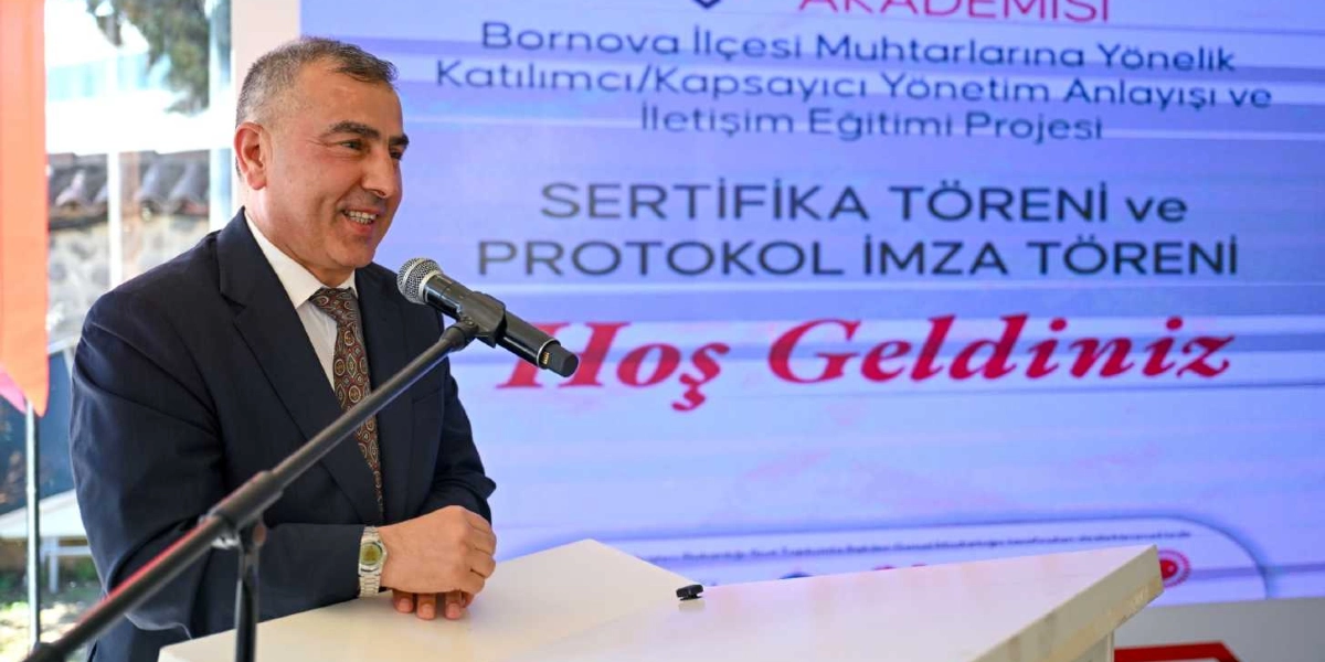 Koyu renk takım elbiseli ve desenli kravatlı bir erkek, önündeki kürsüde bulunan mikrofona konuşma yapıyor. Arka planda yer alan dijital ekranda 