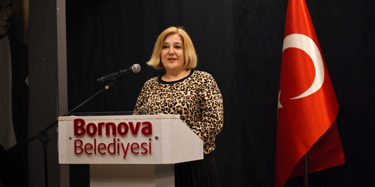 Siyah bir fon önünde, üzerinde Bornova Belediyesi yazılı beyaz bir kürsüde konuşma yapan, sarı saçlı ve leopar desenli bluz giymiş bir kadın. Sağ tarafta Türk bayrağı, sol tarafta ise kısmen görünen bir Atatürk portresi yer alıyor.