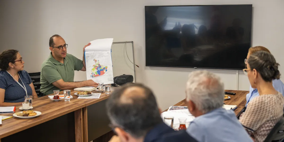 Toplantı masasında oturan bir grup insan, Bornova'nın tarihi dokusunu korumaya yönelik hazırlanan renkli bir imar planı haritasını incelemektedir. Yeşil tişörtlü bir erkek, elinde tuttuğu haritayı masadaki diğer katılımcılara göstermekte ve açıklama yapmaktadır. Masanın üzerinde çay bardakları, su bardakları ve atıştırmalık tabakları yer alırken, arka planda duvara monte edilmiş büyük bir televizyon ekranı bulunmaktadır.