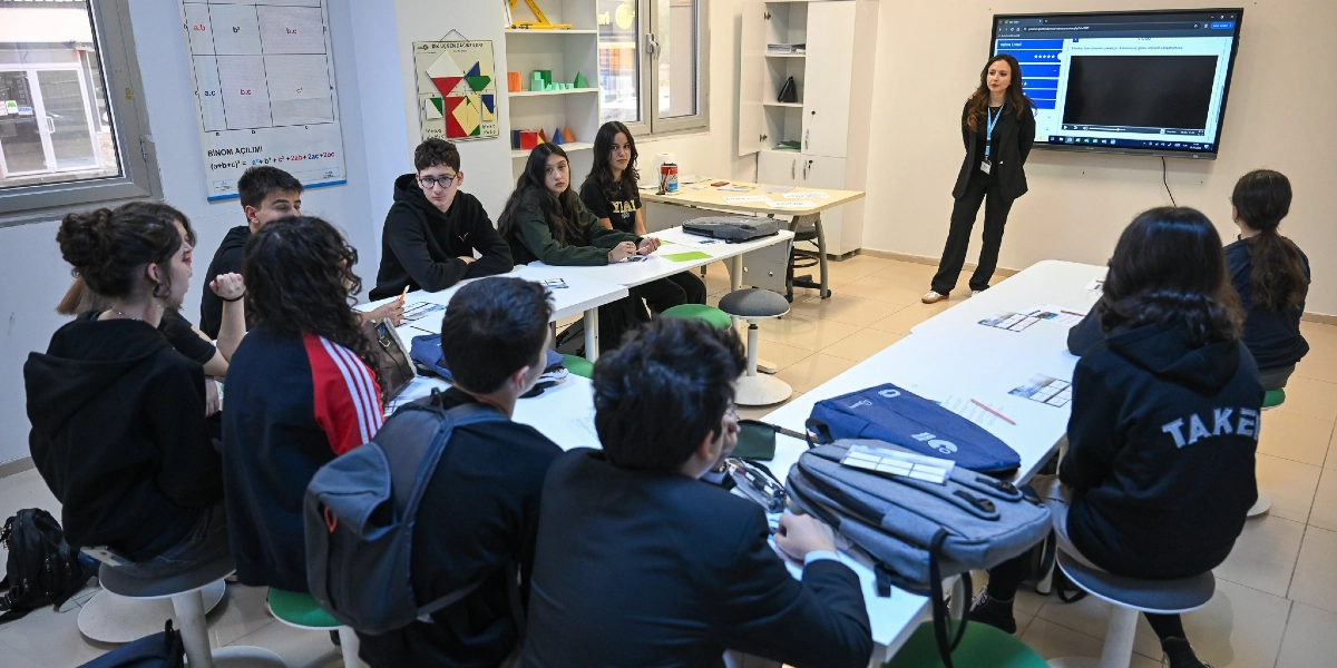 Aydınlık bir sınıfta, beyaz masaların etrafında toplanmış lise çağındaki bir grup öğrenci, ayakta duran kadın eğitmenlerini dinliyor. Eğitmen, duvarda asılı olan büyük bir ekrandan sunum yaparken, öğrenciler masaların üzerindeki ders materyallerini ve dizüstü bilgisayarlarını inceliyor. Sınıfın duvarlarında matematiksel formüllerin ve geometrik şekillerin yer aldığı eğitim posterleri bulunuyor.