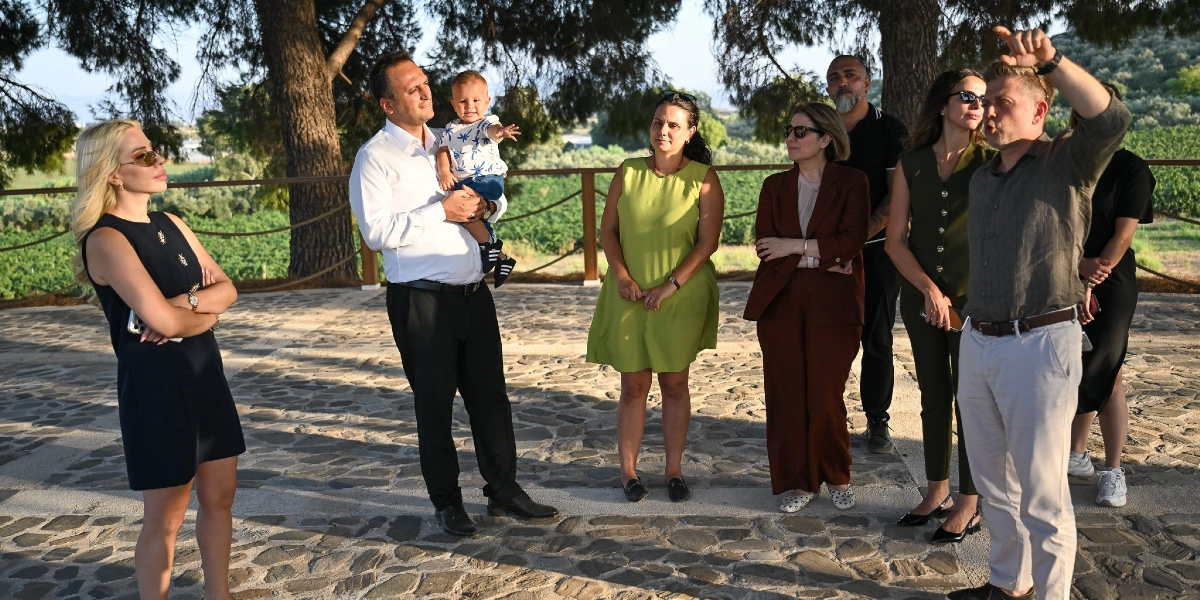Açık havada, taş döşeli bir alanda duran bir grup insan, sağ tarafta duran ve eliyle uzak bir noktayı işaret eden bir adamı dinliyor. Grubun sol tarafında kucağında küçük bir çocuk tutan beyaz gömlekli bir adam, yanında siyah elbiseli bir kadın ve diğer katılımcılar yer alıyor. Arka planda ahşap bir korkuluk ve ağaçların çevrelediği geniş bir yeşil alan bulunuyor.