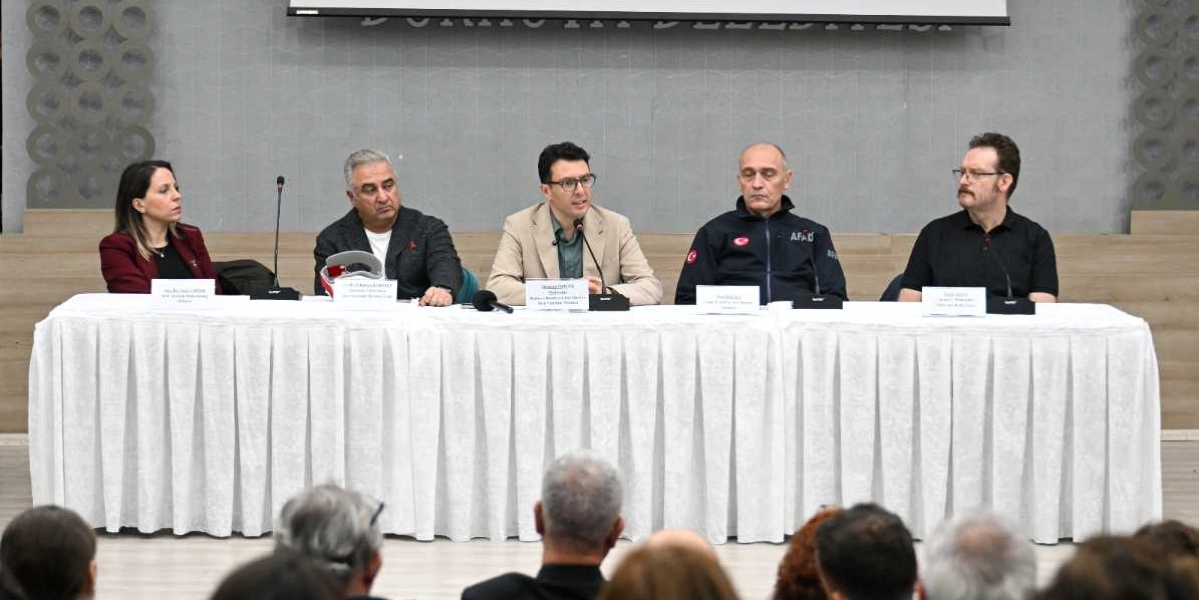 Bornova Belediyesi'nde düzenlenen bir panelde, beyaz örtülü uzun bir masanın arkasında yan yana oturan beş konuşmacı yer alıyor. Soldan sağa doğru; bordo ceketli bir kadın, gri ceketli bir erkek, bej ceketli bir erkek, üzerinde AFAD yazılı lacivert bir ceket giyen bir erkek ve siyah tişörtlü gözlüklü bir erkek oturuyor. Konuşmacıların önünde isimlerinin ve unvanlarının yazılı olduğu küçük beyaz kartlar bulunuyor. Ön planda ise dinleyicilerin başlarının arkası görülüyor.