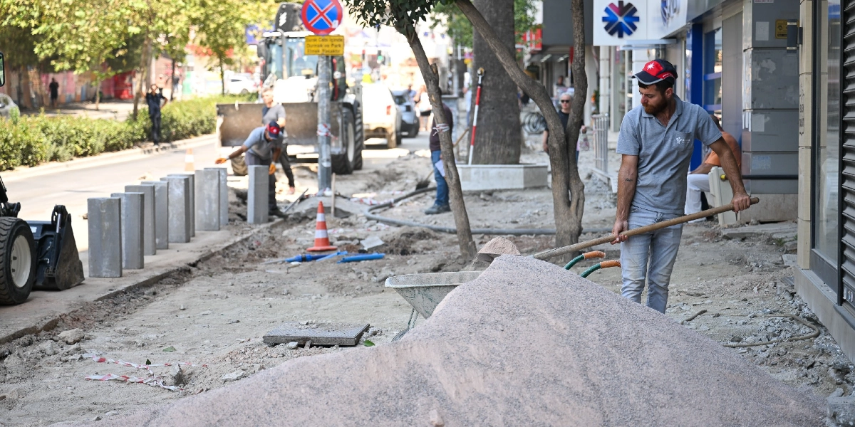 Şehir merkezindeki bir kaldırımda devam eden inşaat çalışmasında, gri tişörtlü ve şapkalı bir işçi elindeki kürekle kum yığınını düzeltiyor. Arka planda başka işçiler, bir iş makinesi, trafik konileri ve kaldırım boyunca dizilmiş beton bariyerler görülüyor.