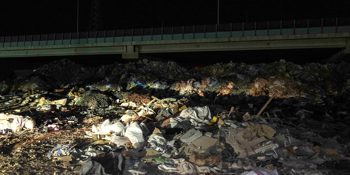 Gece vakti bir otoyol köprüsünün altında birikmiş, inşaat atıkları, molozlar, plastik poşetler ve çeşitli çöplerden oluşan büyük bir yığın.