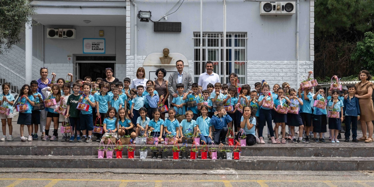 İzmir Bornova Dokuz Eylül İlkokulu binasının önünde, ellerinde içinde küçük bitkiler bulunan hediye paketleri taşıyan çok sayıda öğrenci, öğretmenleri ve okul yöneticileriyle birlikte poz veriyor. Çocukların bir kısmı ellerindeki paketleri okulun girişindeki basamakların önüne dizmiş durumda; arka planda ise okulun giriş kapısı ve üzerinde Atatürk büstü bulunan bir duvar yer alıyor.