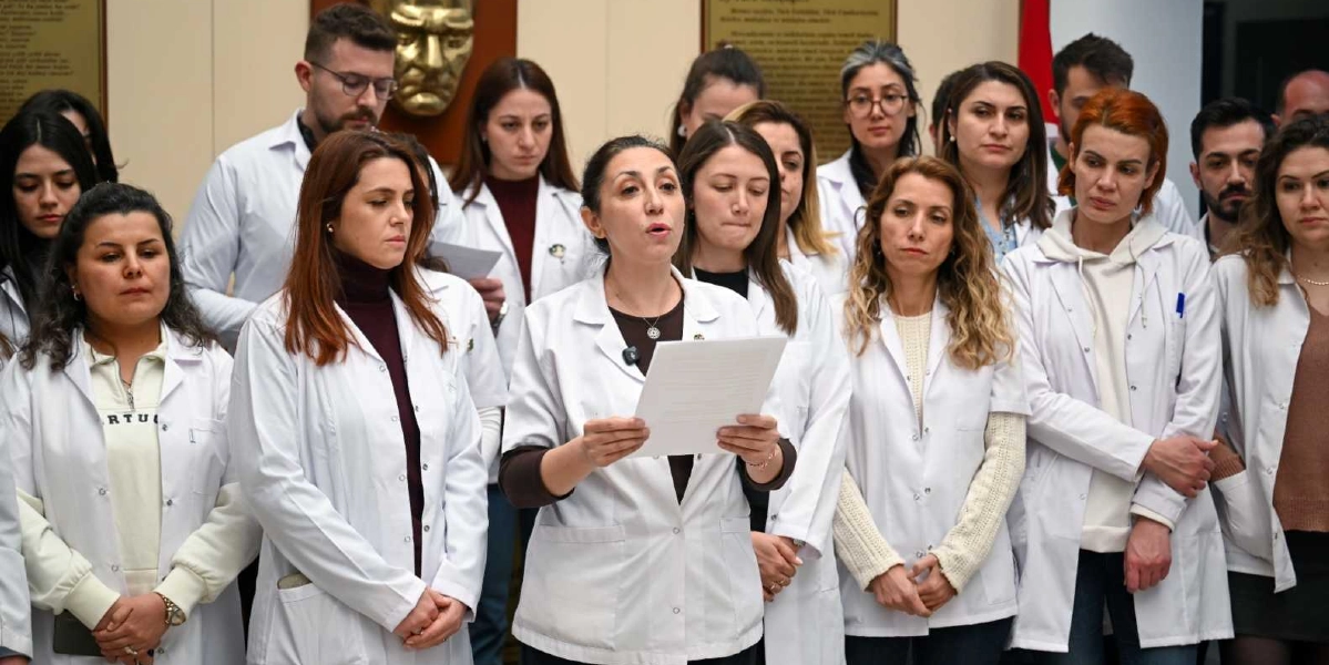Beyaz önlük giymiş bir grup öğretmen, bir basın açıklaması yapmak üzere yan yana duruyor. Grubun önünde yer alan bir kadın öğretmen elindeki metni okurken, diğer öğretmenler ciddi ve kararlı bir ifadeyle onu dinliyor. Arka planda duvarda Atatürk'ün kabartma portresi ve üzerinde yazılı metinler bulunan bir anıt panosu yer alıyor.