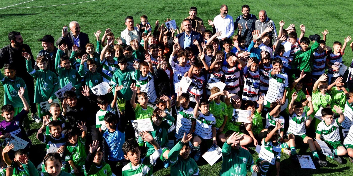 Yeşil bir futbol sahasında, ellerinde katılım belgeleriyle poz veren ve ellerini havaya kaldırarak selamlayan çok sayıda çocuk futbolcu ve yetişkin antrenörler yer alıyor. Farklı renklerde ve desenlerde forma giymiş çocuklar, Atatürk'ü anma turnuvası kapsamında bir araya gelmiş, neşeli bir şekilde objektife bakıyorlar. Arka planda ayakta duran yetişkinler de çocukların başarısını kutluyor.