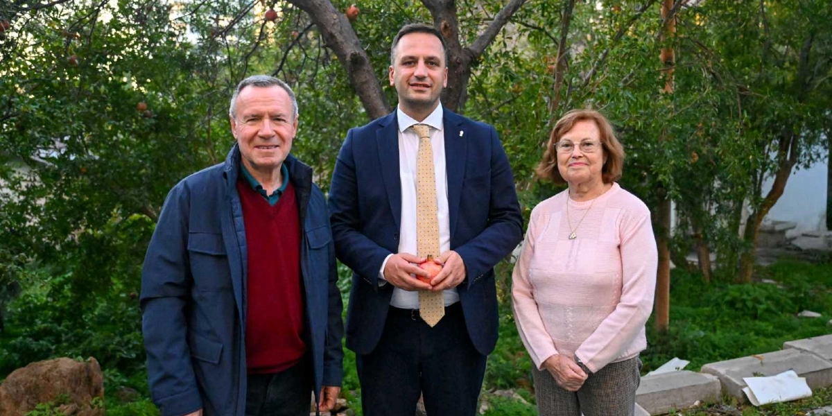 Açık havada, ağaçlık bir alanda yan yana duran üç kişi. Ortada lacivert takım elbiseli, bej kravatlı bir erkek ellerinde bir nar tutuyor. Onun solunda bordo kazak ve lacivert ceket giyen bir erkek, sağında ise açık pembe bir kazak giyen gözlüklü bir kadın yer alıyor.