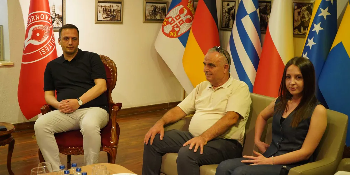 Bornova Belediyesi'ni ziyaret eden Makedon heyeti, toplantı odasında yan yana otururken görülüyor. Solda, üzerinde Bornova Belediyesi logosu bulunan kırmızı bir bayrağın önünde, siyah tişörtlü ve açık renk pantolonlu bir erkek tekli koltukta oturuyor. Yanında, arkalarında Sırbistan, Almanya, Yunanistan, Polonya ve Bosna-Hersek bayraklarının bulunduğu bir koltukta, krem rengi polo yaka tişörtlü bir erkek ve yanında lacivert yelekli bir kadın oturuyor. Arka planda duvarda çerçeveli fotoğraflar asılı.