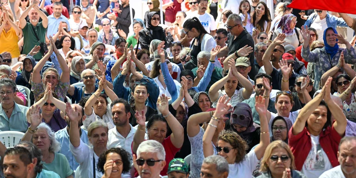 Açık havada düzenlenen bir etkinlikte, ellerini havaya kaldırarak alkışlayan ve neşeli görünen kalabalık bir insan grubu. Farklı yaş ve giyim tarzlarından oluşan katılımcılar, güneşli bir havada bir arada bulunuyor.