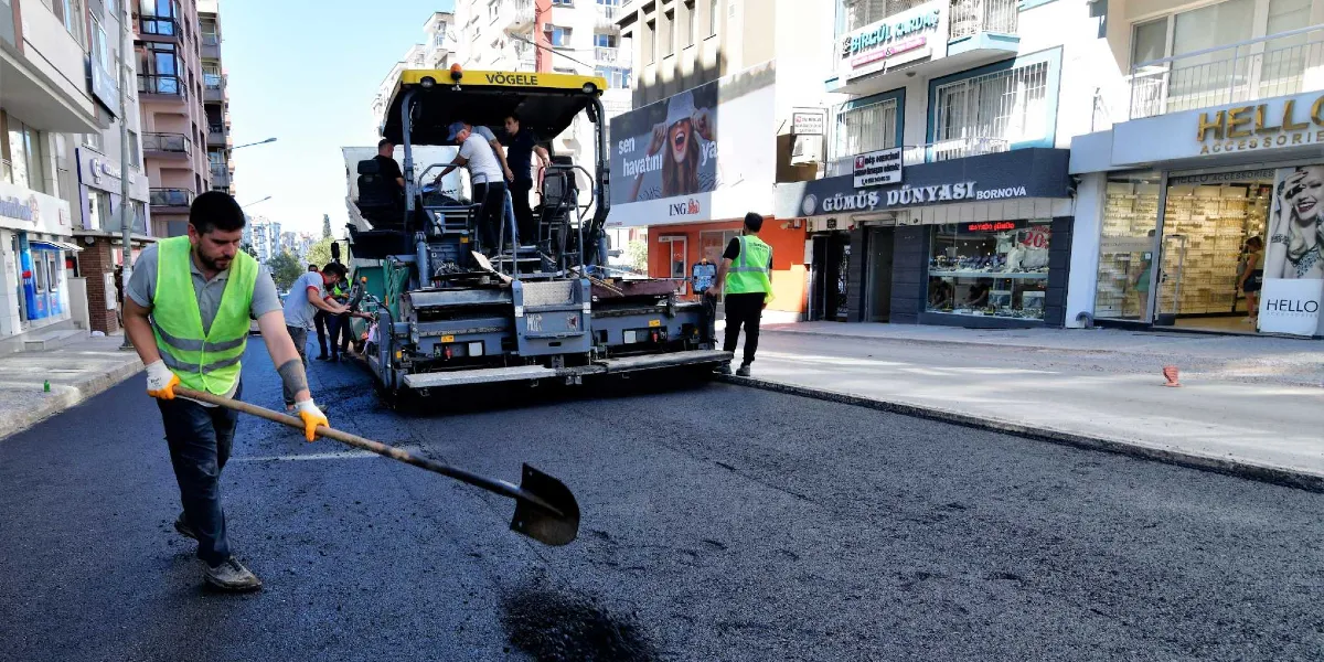 Bornova'da bir cadde üzerinde işçiler tarafından yürütülen asfalt serim çalışması. Ön planda sarı reflektörlü yelek giyen bir işçi elindeki kürekle asfaltı düzeltirken, arka planda Vögele marka bir asfalt serme makinesi ve diğer görevliler yer alıyor. Çevredeki dükkanların tabelaları ve cadde üzerindeki hareketlilik görülüyor.