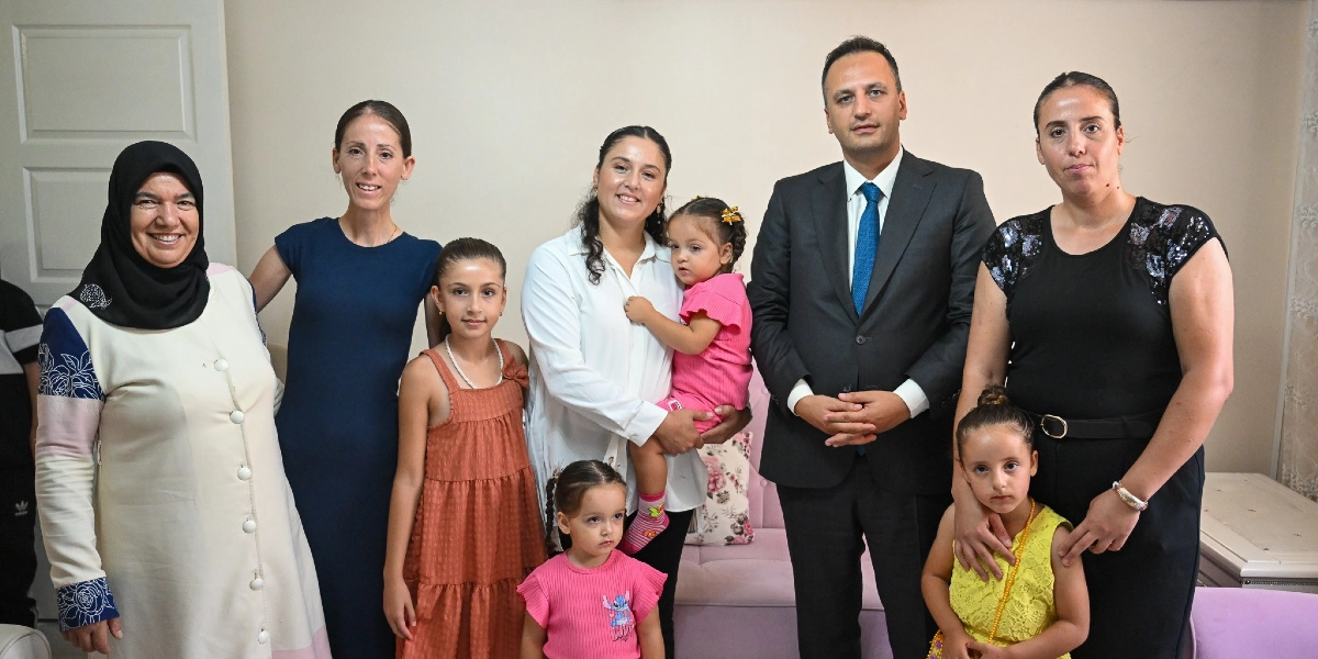 İç mekan bir odada bir araya gelmiş bir grup insan; solda başörtülü, krem rengi uzun bir kıyafet giyen bir kadın, yanında lacivert elbiseli bir kadın, önlerinde kiremit rengi elbiseli bir kız çocuğu, ortada beyaz gömlekli bir kadın kucağında pembe kıyafetli küçük bir kız çocuğu, onun önünde pembe tişörtlü küçük bir kız çocuğu, sağ tarafta lacivert takım elbiseli ve mavi kravatlı bir erkek, yanında siyah bluzlu bir kadın ve onun önünde sarı elbiseli bir kız çocuğu yan yana durarak gülümsüyor.