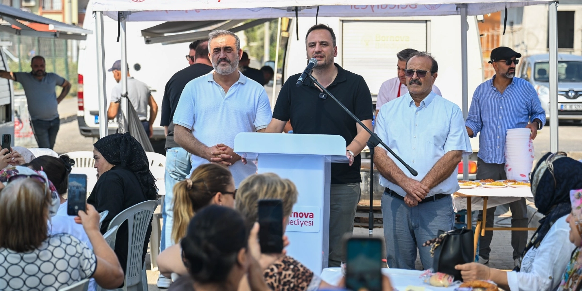 Bornova Belediyesi'ne ait bir çadırın altında, kürsüde konuşma yapan bir yetkili ve yanında duran iki kişi, açık havada düzenlenen bir etkinlikte katılımcılara hitap ediyor. Kürsüdeki kişi mikrofonla konuşurken, yanında beyaz gömlekli iki erkek eşlik ediyor. Etkinlik alanında oturan katılımcılar, konuşmayı cep telefonlarıyla kaydediyor ve dinliyor. Arka planda Bornova Belediyesi logolu bir araç ve çevredeki insanlar görülüyor.