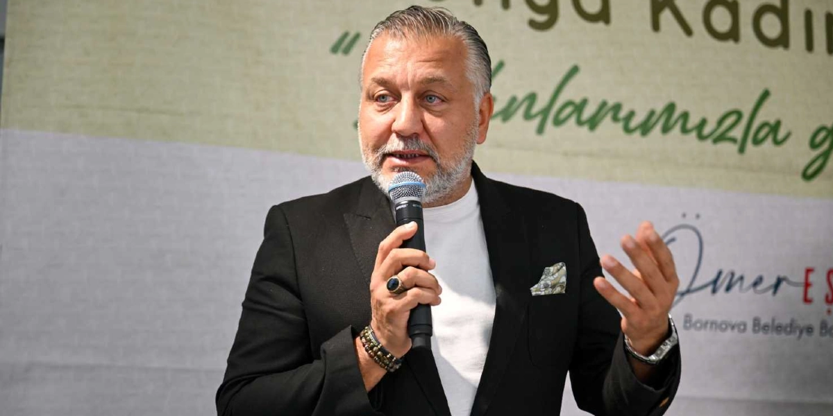 Bornova Belediye Başkanı Ömer Eşki, siyah ceket ve beyaz tişört giymiş bir şekilde elinde mikrofonla konuşma yapıyor. Arka planda üzerinde 