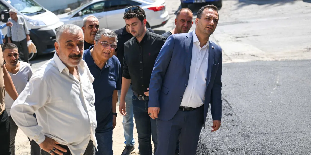 Lacivert takım elbiseli bir erkek, yanında bulunan bir grup erkekle birlikte yeni dökülmüş asfalt çalışmasını inceliyor. Grup, açık havada, asfaltlanmış bir yolun kenarında duruyor ve dikkatlerini yola vermiş durumda.