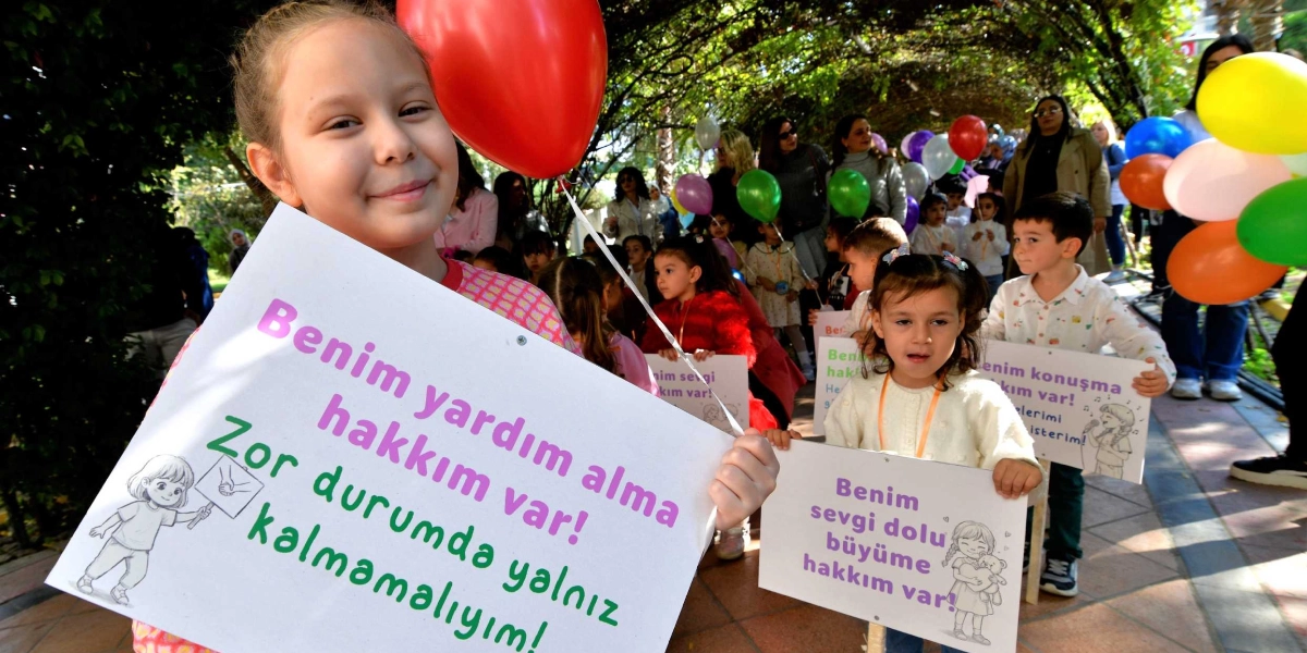 Açık havada, yeşilliklerle kaplı bir yolda yürüyen çocuklar, ellerinde çocuk haklarıyla ilgili sloganların yazılı olduğu pankartlar ve renkli balonlar taşıyor. Ön planda, elinde kırmızı bir balon tutan ve üzerinde 