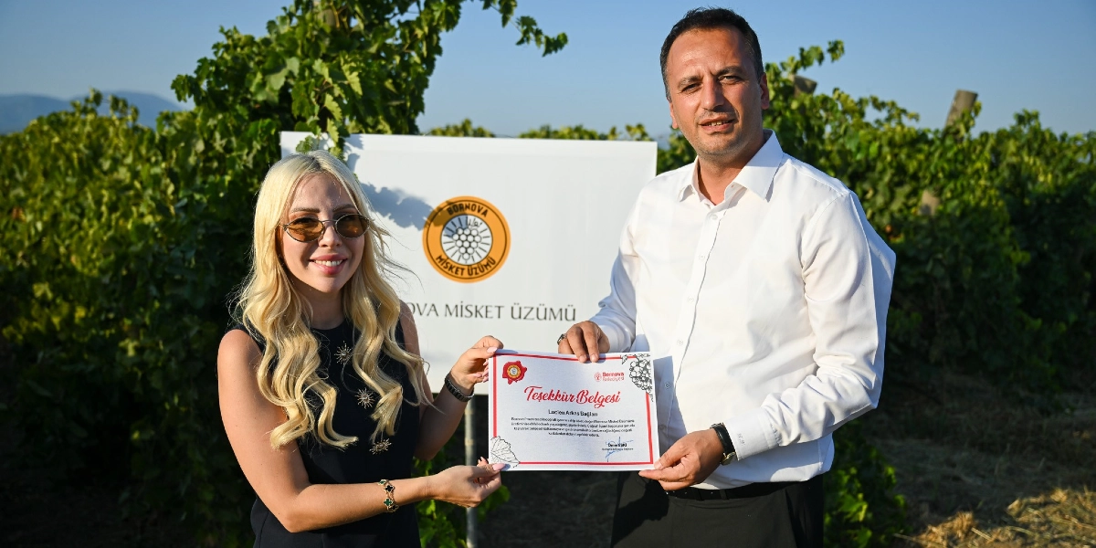 Arka planda üzüm bağlarının bulunduğu bir alanda, sarı saçlı bir kadın ve beyaz gömlekli bir erkek, üzerinde 