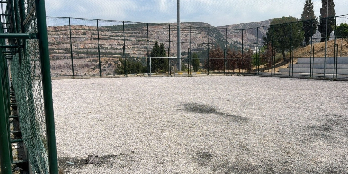 Etrafı yüksek tel örgülerle çevrili, zemini tamamen çakıl taşlarıyla kaplı, içinde tek bir futbol kalesi bulunan açık hava spor sahası. Sahanın arka planında kayalık bir tepe ve ağaçlar yer alıyor.