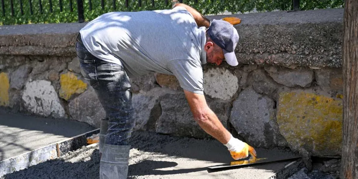 Gri tişörtlü, şapkalı ve iş eldivenli bir işçi, taş duvarın önündeki taze dökülmüş betonu mala ile düzeltiyor.