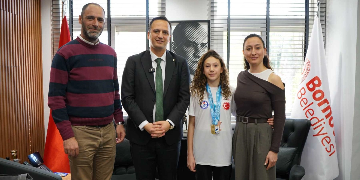 Bornova Belediye Başkanı, yanında madalyalı genç bir sporcu ve iki yetişkin ile birlikte ofisinde poz veriyor. Arka planda Türk bayrağı ve üzerinde Bornova Belediyesi yazılı bir flama bulunuyor.