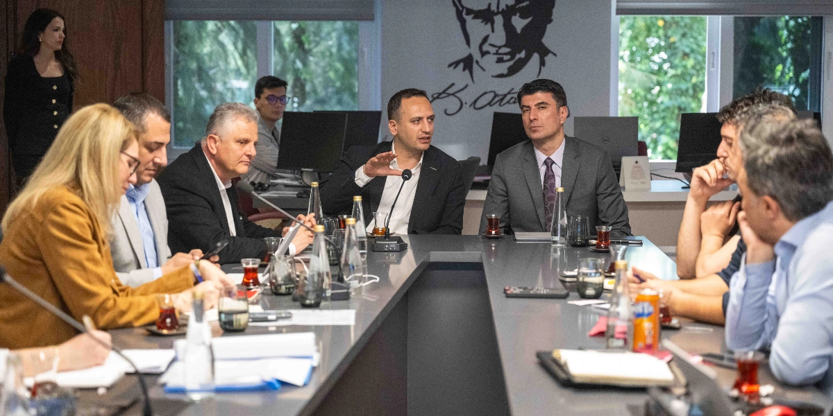 Modern bir ofis ortamında, uzun bir toplantı masasının etrafında toplanmış, iş kıyafetleri içindeki bir grup insan toplantı yapıyor. Masanın merkezinde konuşan bir kişi el hareketleriyle bir şeyler anlatırken, diğer katılımcılar onu dikkatle dinliyor. Masanın üzerinde çay bardakları, su şişeleri, not defterleri ve mikrofonlar bulunuyor. Arka plandaki duvarda Mustafa Kemal Atatürk'ün silüeti ve imzası yer alıyor.