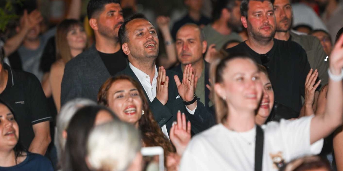 Açık havada düzenlenen bir etkinlikte, ellerini çırparak ve neşeyle şarkıya eşlik ederek eğlenen kalabalık bir grup insan. Ön planda beyaz tişörtlü bir kadın ve koyu renk ceketli bir adam dikkat çekerken, arka planda farklı yaş gruplarından insanlar coşkuyla etkinliğe katılıyor.