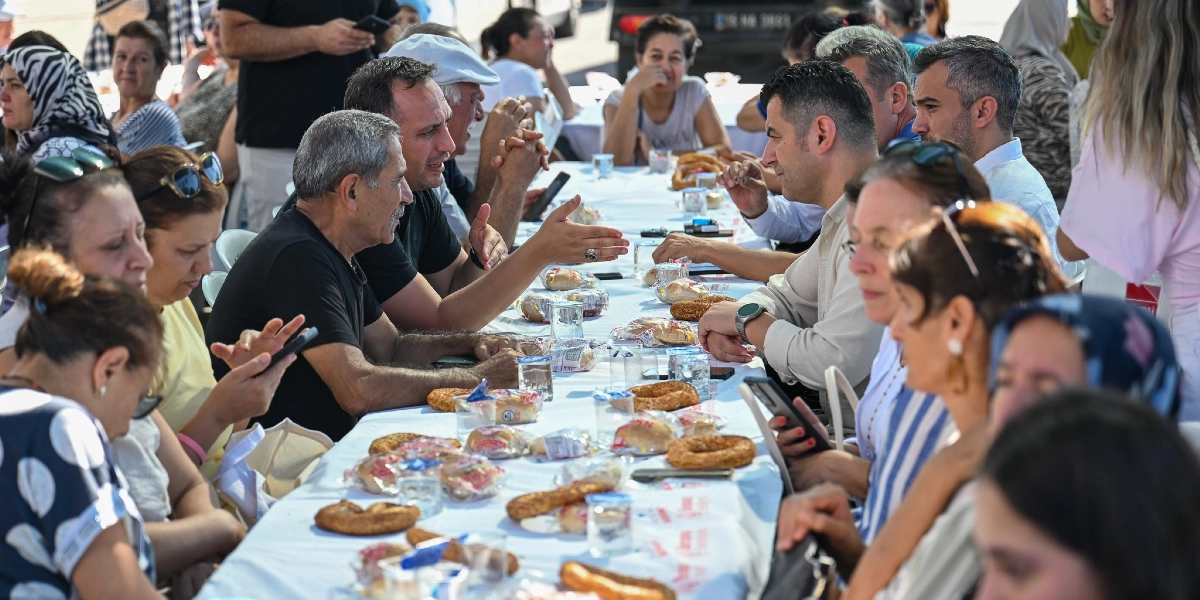 Açık havada uzun bir masada bir araya gelen grup, beyaz örtülü masada simit ve çay eşliğinde sohbet ediyor. Masanın başında oturan Başkan Eşki, yanındaki katılımcılarla konuşurken, çevresindeki kadın üreticiler ve diğer katılımcılar masada oturuyor. Bazı katılımcılar telefonlarıyla ilgilenirken, bazıları ise karşılıklı sohbet ediyor.