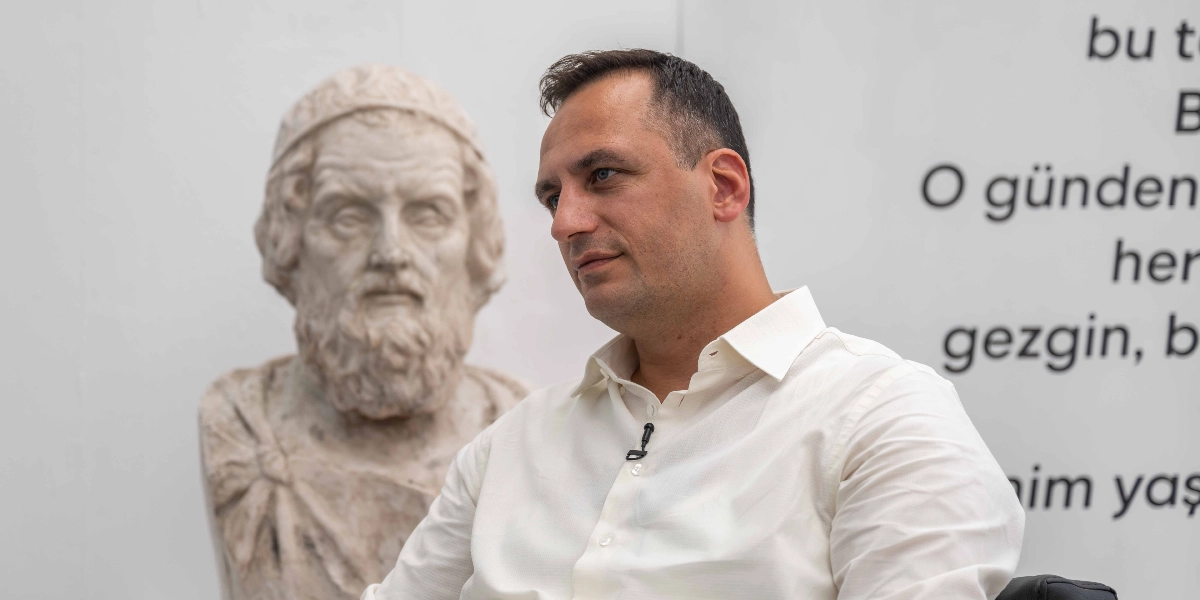 Beyaz gömlekli, yakasında yaka mikrofonu bulunan bir erkek, arka planda yer alan antik bir büstün önünde sağ tarafa doğru bakıyor. Arka plandaki beyaz duvarda kısmen görünen Türkçe metinler yer alıyor.