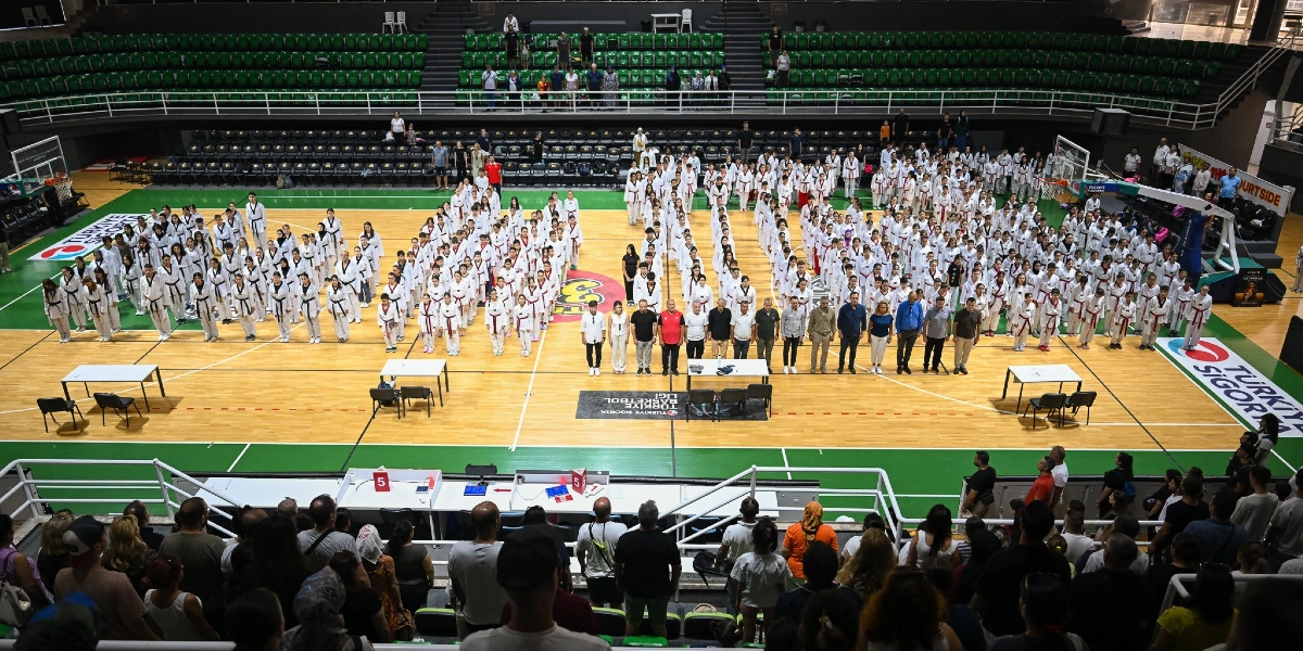 Kapalı bir spor salonunda, parke zemin üzerinde düzenli sıralar halinde dizilmiş, beyaz tekvando kıyafetleri giyen çok sayıda sporcu adayı ve önlerinde duran bir grup yetkili görülüyor. Sporcuların bir kısmı renkli kuşaklar takmış durumda. Salonun tribünlerinde ise izleyiciler oturuyor. Parke zeminin üzerinde Türkiye Sigorta Basketbol Ligi logoları yer alıyor.