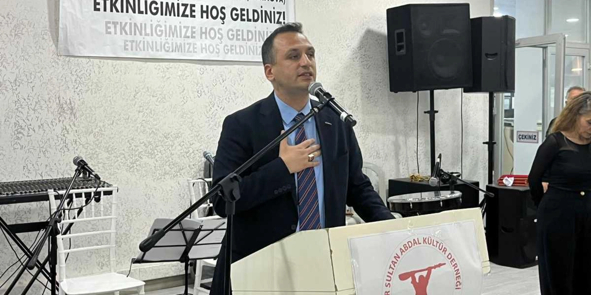 Kürsüde konuşma yapan, lacivert takım elbiseli ve çizgili kravatlı bir erkek, sağ elini göğsüne koymuş şekilde mikrofon başında duruyor. Kürsünün ön yüzünde Pir Sultan Abdal Kültür Derneği'nin logosu yer alıyor. Arka planda üzerinde 