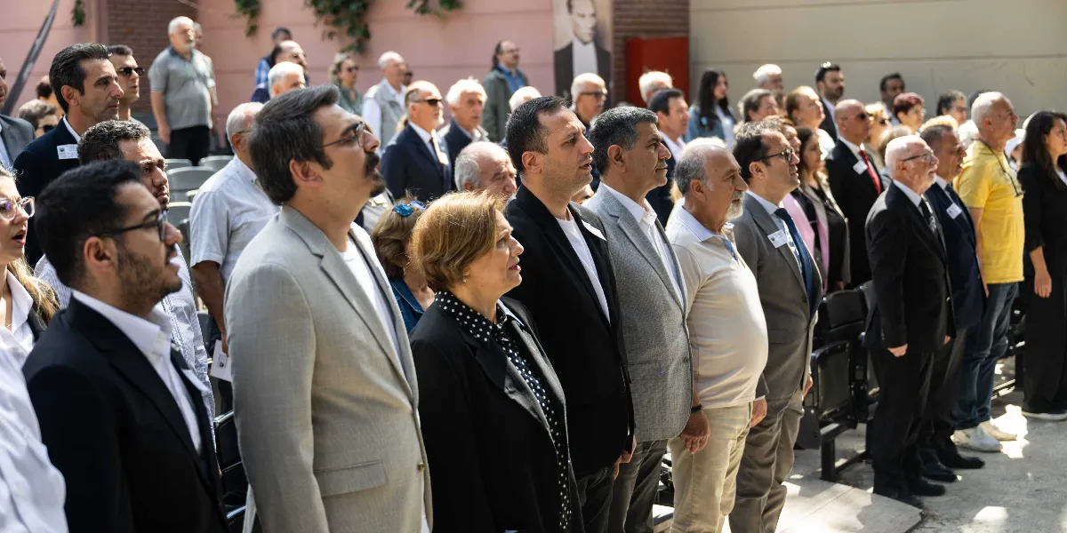 Atatürkçü Düşünce Derneği Genel Kurulu'nda bir araya gelen kalabalık bir grup, saygı duruşu veya İstiklal Marşı sırasında ciddiyetle ayakta duruyor. Ön planda, takım elbiseli ve günlük kıyafetli kadın ve erkeklerden oluşan katılımcılar, hafifçe yukarıya veya karşıya bakarak bir tören anını paylaşıyor. Arka planda ise duvar üzerinde Mustafa Kemal Atatürk'ün bir portresi yer alıyor.