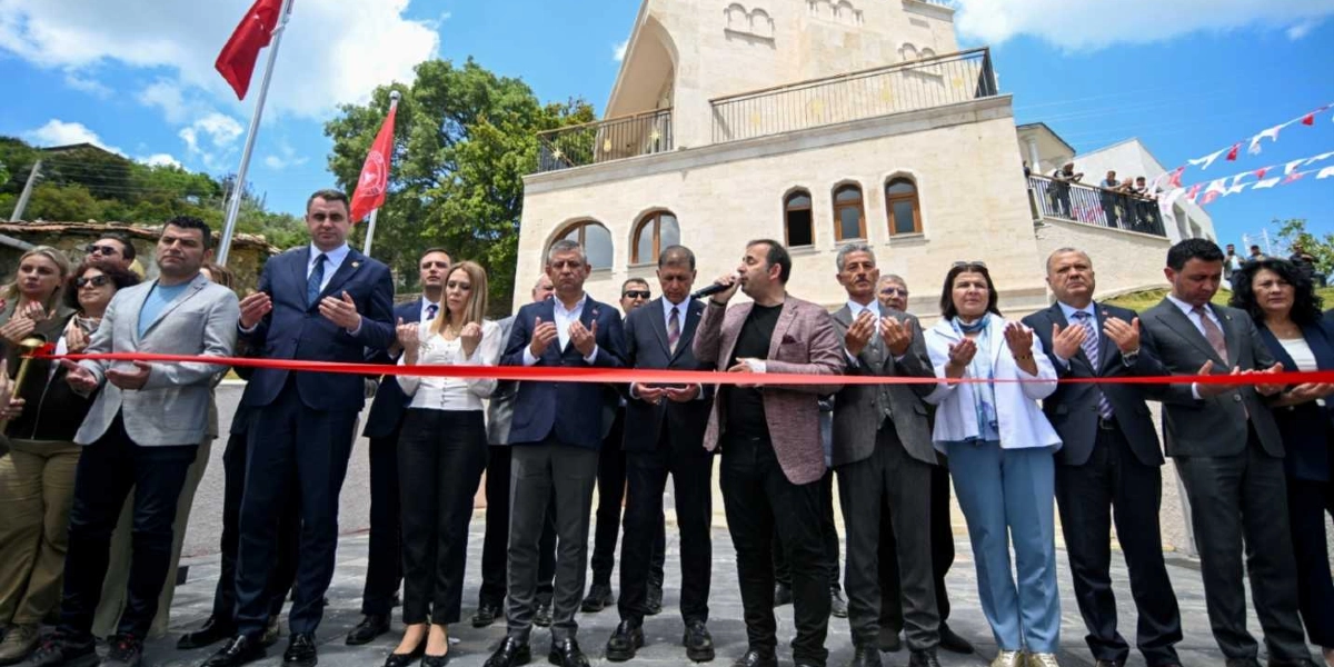 CHP Genel Başkanı Özgür Özel ve beraberindeki heyet, Bornova'da yeni bir tesisin açılış töreninde ellerini açarak dua ediyor. Açılış kurdelesinin önünde yan yana dizilmiş grup, takım elbiseli erkekler ve şık giyimli kadınlardan oluşuyor. Arka planda taş mimariye sahip, kemerli pencereleri olan iki katlı bir yapı ve gökyüzünde dalgalanan Türk bayrakları ile parti bayrakları görülüyor.
