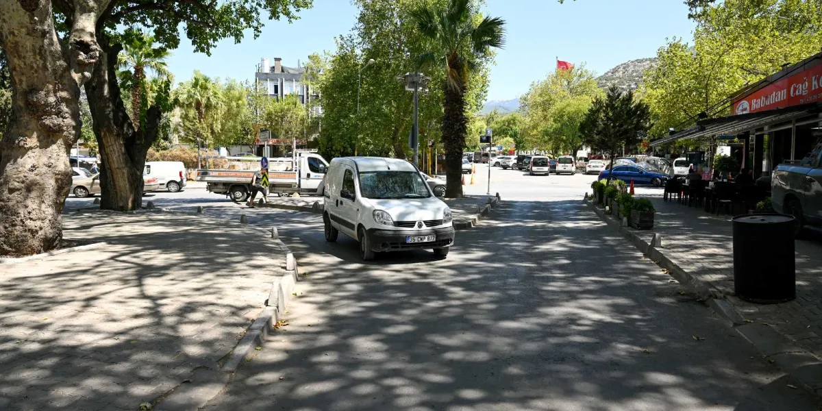 Geniş ağaçların gölgelediği, sağ tarafında Babadan Cağ Kebap restoranının bulunduğu Pınar Caddesi'nde, beyaz bir hafif ticari araç ilerliyor. Yolun kenarlarında park etmiş araçlar, arka planda ise ağaçların arasında görünen bir Türk bayrağı ve dağ manzarası yer alıyor.