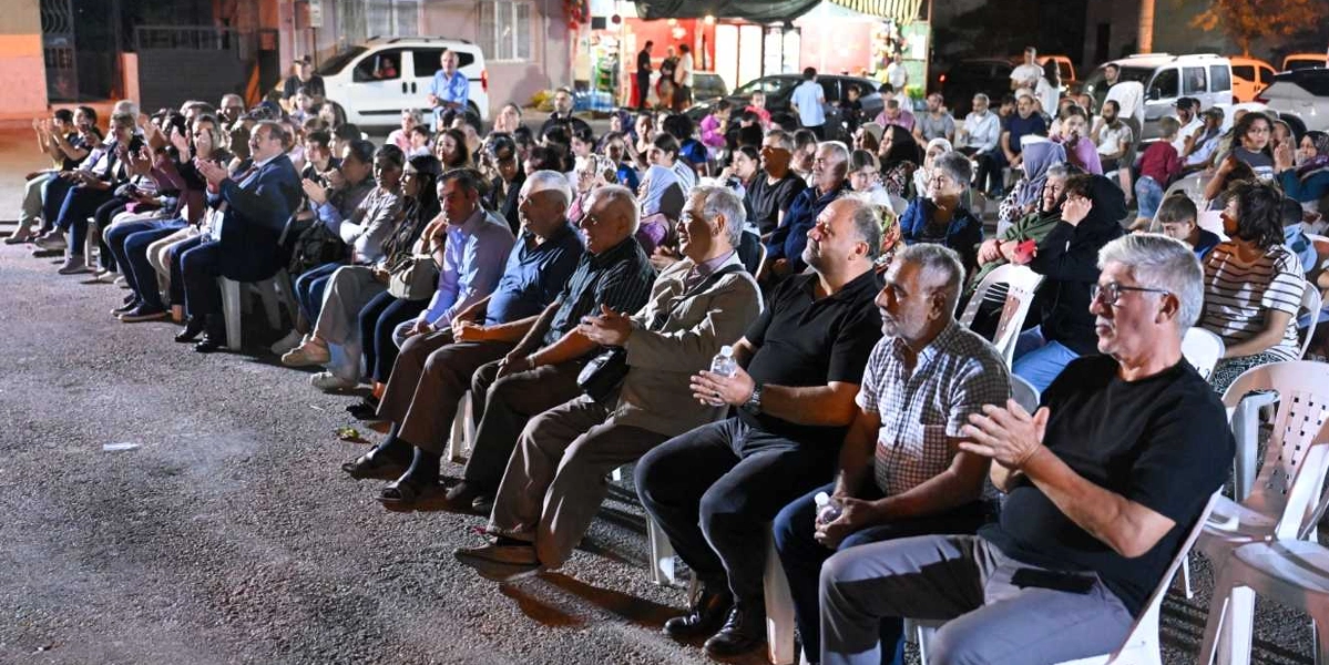 Açık havada, akşam saatlerinde düzenlenen bir etkinlikte beyaz plastik sandalyelerde yan yana oturmuş, sahneyi izleyen ve alkışlayan kalabalık bir izleyici grubu. Ön planda, siyah tişörtlü, gri saçlı ve gözlüklü bir adamın yanı sıra farklı yaş gruplarından kadın ve erkekler dikkatle sahneye bakıyor. Arka planda park halindeki araçlar ve etkinliğin yapıldığı alanın ışıklandırılmış çevresi görülüyor.