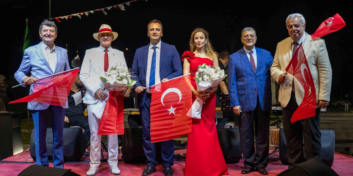 Gece vakti açık hava sahnesinde yan yana duran altı kişilik grup, ellerinde Türk bayrakları ve çiçek buketleriyle 30 Ağustos Zafer Bayramı resepsiyonunda poz veriyor. Soldan sağa doğru; açık mavi ceketli bir erkek, beyaz takım elbiseli ve şapkalı bir erkek, lacivert takım elbiseli bir erkek, kırmızı uzun bir elbise giyen bir kadın, mavi ekose ceketli bir erkek ve bej renkli ceket giyen bir erkek yer alıyor. Arka planda müzik aletleri ve sahne ekipmanları görülüyor.