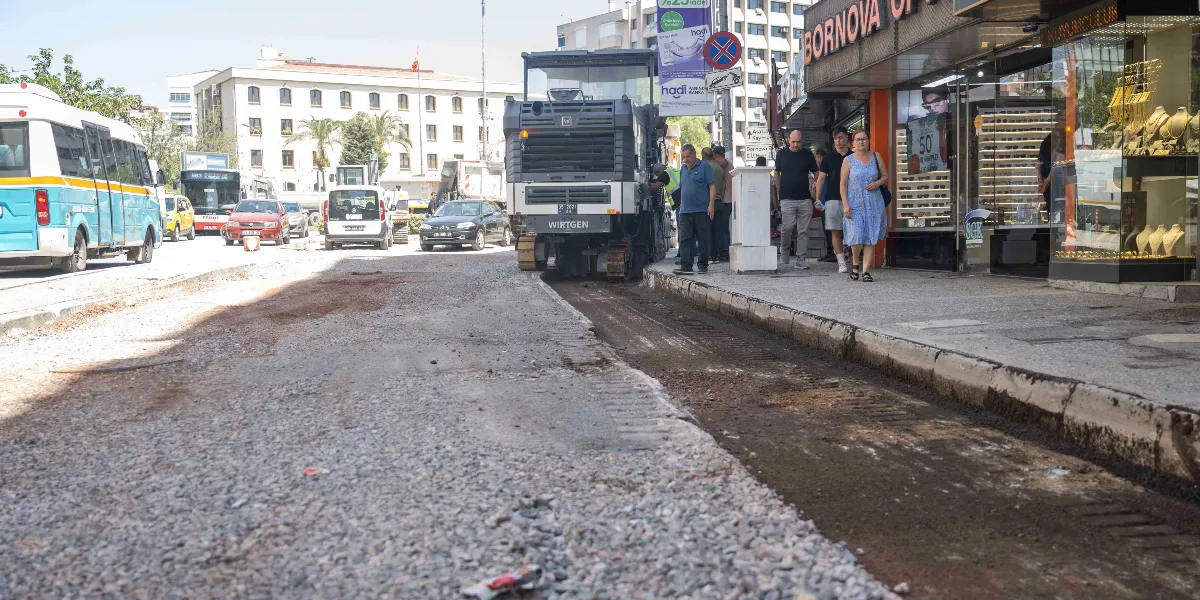 Mustafa Kemal Caddesi'nde devam eden yol yenileme çalışmaları kapsamında, asfalt kazıma makinesi caddenin bir şeridinde çalışırken, yayalar kaldırımda yürümeye devam ediyor. Yolun bir kısmı kazınmış ve çakıllı bir zemin görünürken, arka planda trafik akışı ve çevredeki dükkanlar yer alıyor.